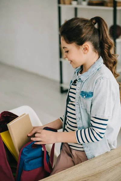 Entzückendes Kind, das Bücher in den Rucksack packt, während es zu Hause am Schreibtisch steht — Stockfoto