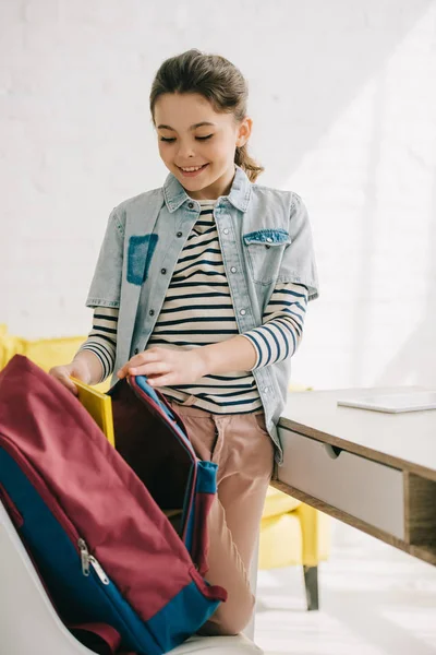 Entzückend lächelndes Kind packt Rucksack, während es am Schreibtisch zu Hause steht — Stockfoto