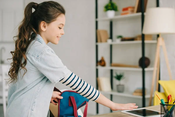 Linda mochila de embalaje escolar mientras está de pie cerca del escritorio con tableta digital con pantalla en blanco en casa - foto de stock