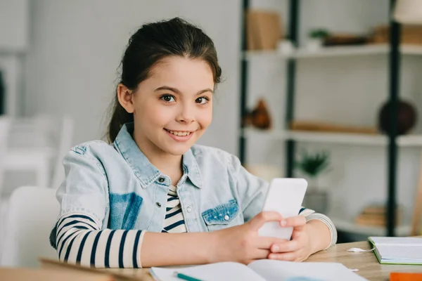 Criança adorável usando smartphone, sorrindo e olhando para a câmera — Fotografia de Stock