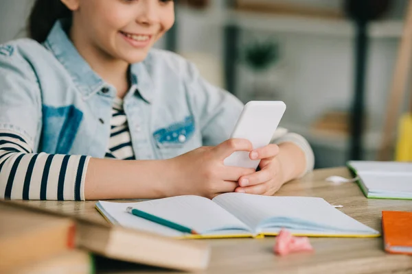 Vista parcial de un niño sonriente usando un teléfono inteligente mientras está sentado en el escritorio y haciendo trabajo escolar en casa - foto de stock