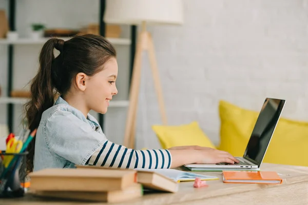 Vista laterale del bambino adorabile che usa il computer portatile mentre è seduto alla scrivania con i libri e fa i compiti — Foto stock