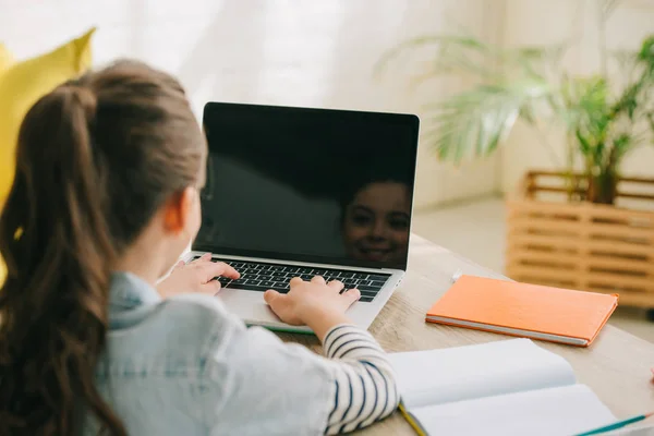 Back view of cute schoolchild using laptop while sitting at desk with copy books — Stock Photo