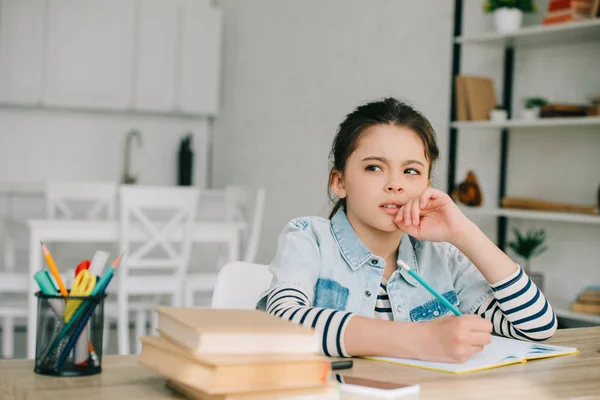 Вдумчивый ребенок смотрит в сторону, сидя за столом и делать домашнее задание — стоковое фото