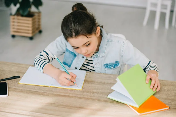 Criança atenta que escreve no livro da cópia e que olha no livro ao fazer o dever de casa — Fotografia de Stock