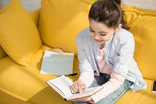 Мила усміхнена дитина читає книгу, сидячи на жовтому дивані вдома — Stock Photo