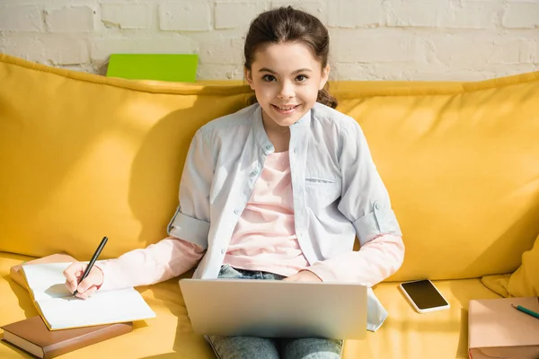 Adorable enfant écrit dans un ordinateur portable et en utilisant un ordinateur portable tout en étant assis sur un canapé jaune près de livres et smartphone avec écran blanc — Photo de stock