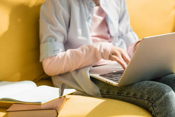 Vista parziale del bambino che usa il computer portatile seduto sul divano giallo vicino al libro e copia il libro — Foto stock