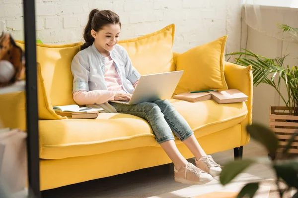 Adorable child using laptop while sitting on yellow sofa near books at home — Stock Photo