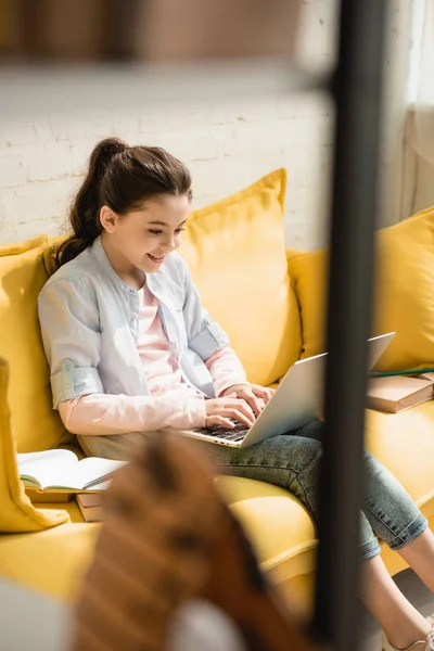 Selektiver Fokus des lächelnden Kindes mit Laptop, während es zu Hause auf dem Sofa neben Büchern sitzt — Stockfoto