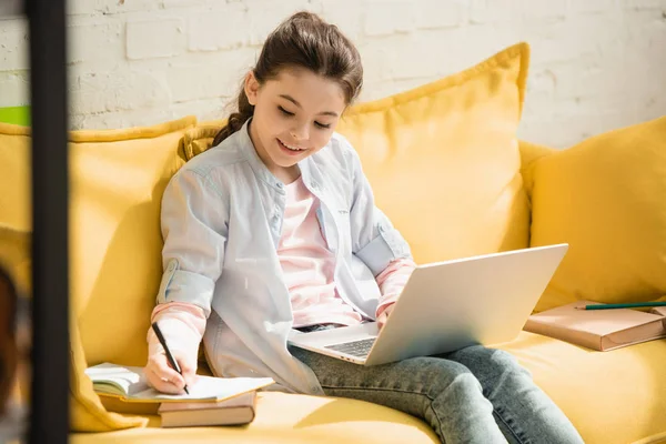 Улыбающийся внимательный ребенок пишет в копировальной книге и с помощью ноутбука, сидя на диване дома — стоковое фото
