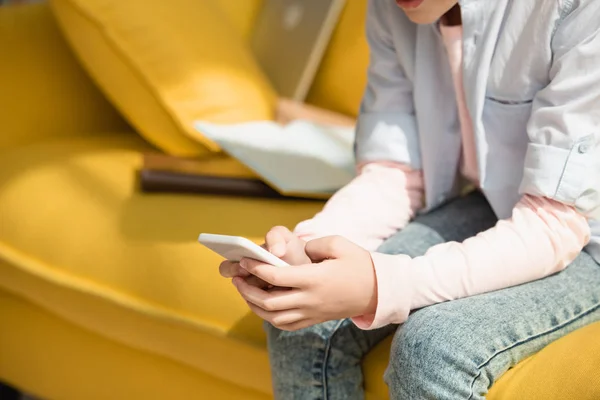 Vista recortada del niño usando el teléfono inteligente mientras está sentado en el sofá en casa - foto de stock