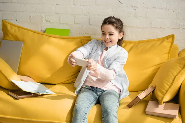 Cheerful child taking selfie while sitting on yellow sofa near books at home — Stock Photo