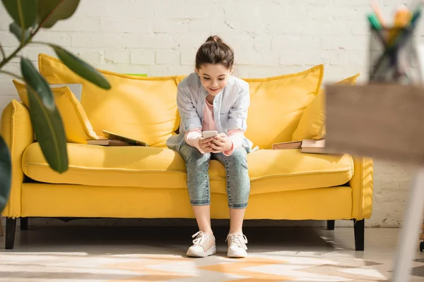 Foco seletivo da criança bonito usando smartphone enquanto sentado no sofá amarelo em casa — Fotografia de Stock