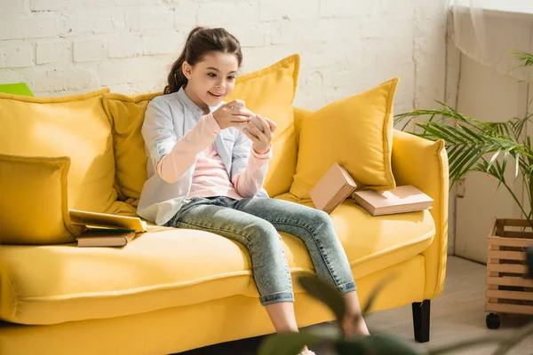 Entzückend lächelndes Kind sitzt auf Sofa neben Büchern und benutzt Smartphone — Stockfoto