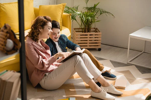 Cheerful mother and son sitting on floor at home and reading book together — Stock Photo