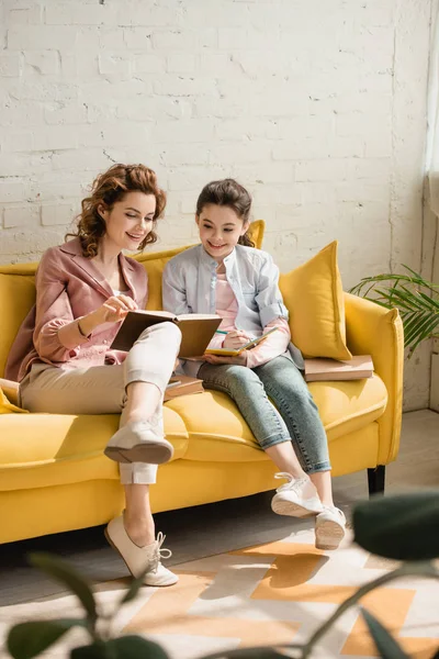 Alegre madre e hija sentadas en un sofá amarillo y leyendo un libro juntas - foto de stock