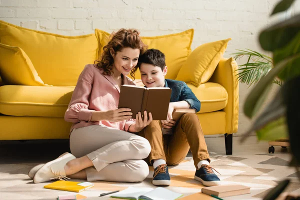 Улыбающиеся мать и сын сидят на полу возле желтого дивана и вместе читают книгу — стоковое фото