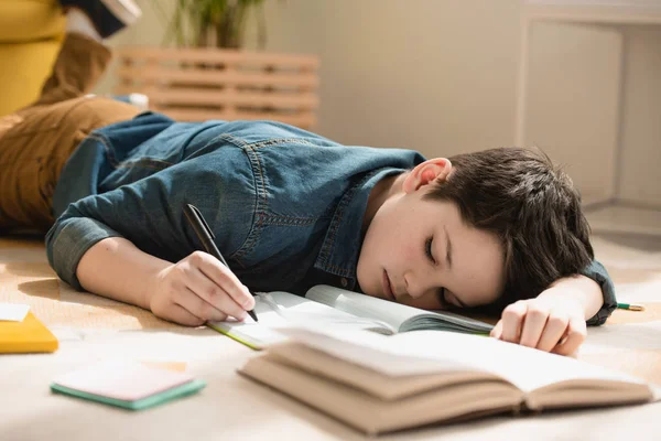 Niño cansado acostado en el suelo y escribiendo en el libro de copia mientras hace el trabajo escolar en casa - foto de stock