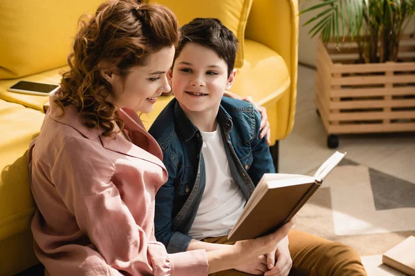 Щаслива мати обіймає чарівного сина, сидячи на підлозі і читаючи книгу разом — стокове фото