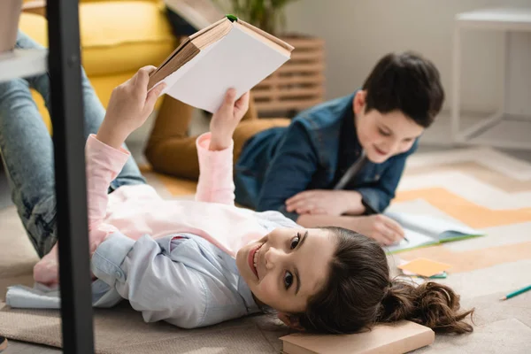 Messa a fuoco selettiva di bambino allegro che si trova sul pavimento con libro vicino a fratello che scrive in notebook — Foto stock