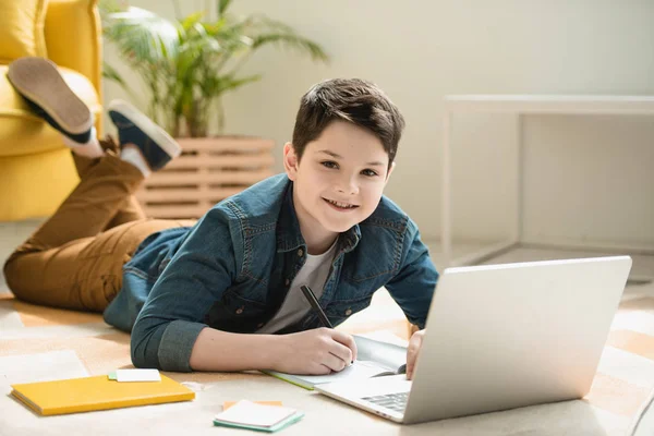 Cheerful boy lying on floor, writing in copy book and using laptop while smiling at camera — Stock Photo