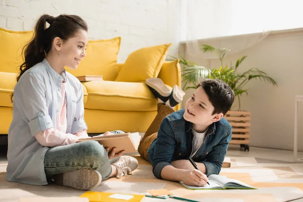 Smiling boy lying on floor and writing in copy book near sister sitting with book — Stock Photo