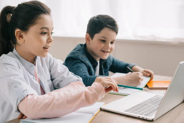 Adorabili bambini seduti alla scrivania, che scrivono in libri di copia e utilizzano il computer portatile insieme — Foto stock