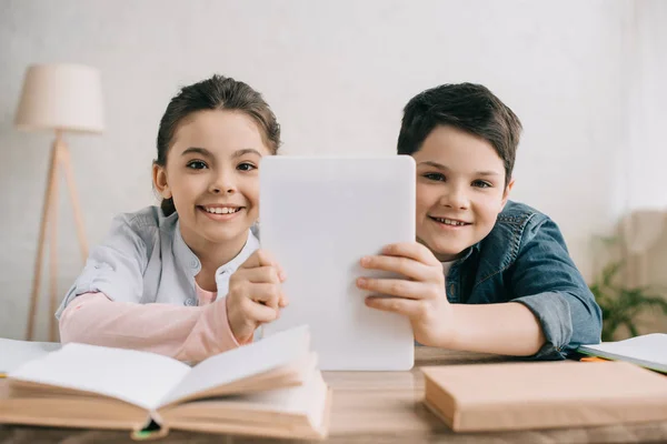 Cheerful children holding digital tablet and looking at camera while sitting at table with books together — Stock Photo