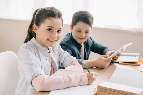 Foyer sélectif de sourire écriture enfant dans le carnet et en regardant la caméra tout en faisant des devoirs avec le frère joyeux — Photo de stock