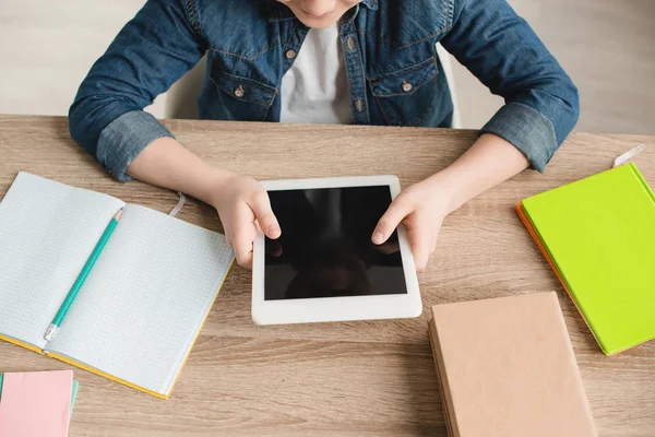 Vista recortada de niño sosteniendo tableta digital con pantalla en blanco mientras está sentado en el escritorio con cuadernos - foto de stock