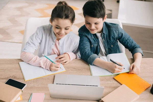 Sonriente hermano y hermana escribiendo en cuadernos y usando el ordenador portátil juntos - foto de stock