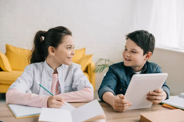 Smiling boy using digital tablet while sitting at desk near sister writing in notebook — Stock Photo