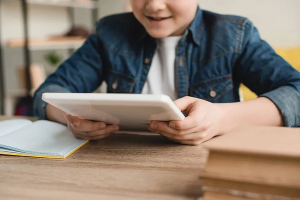 Teilaufnahme eines lächelnden Jungen, der ein digitales Tablet benutzt, während er zu Hause mit Büchern am Schreibtisch sitzt — Stockfoto