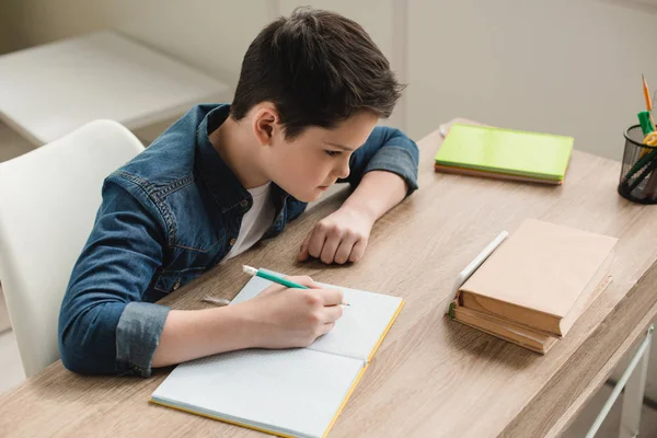 Vista de ángulo alto de niño atento escribiendo en el libro de copia mientras que hace el trabajo escolar en casa - foto de stock