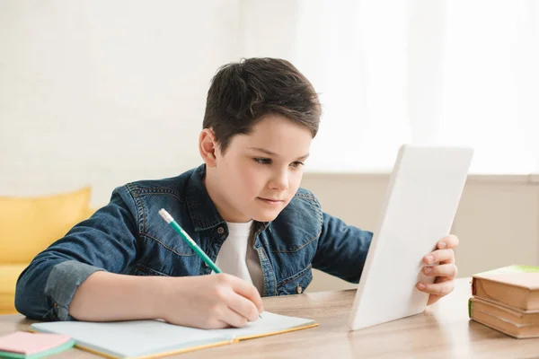 Niño concentrado escribiendo en el cuaderno y usando la tabla digital mientras que hace el trabajo escolar en el país - foto de stock