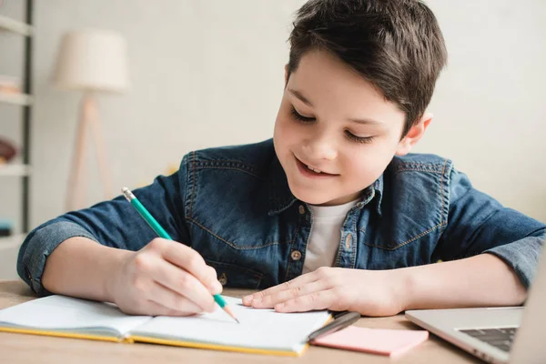 Fröhlicher Junge schreibt in Notizbuch, während er am Schreibtisch sitzt und Hausaufgaben macht — Stockfoto