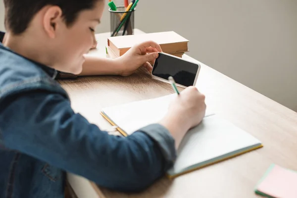 Selective focus of boy writing in notebook and holding smartphone with blank screen — Stock Photo