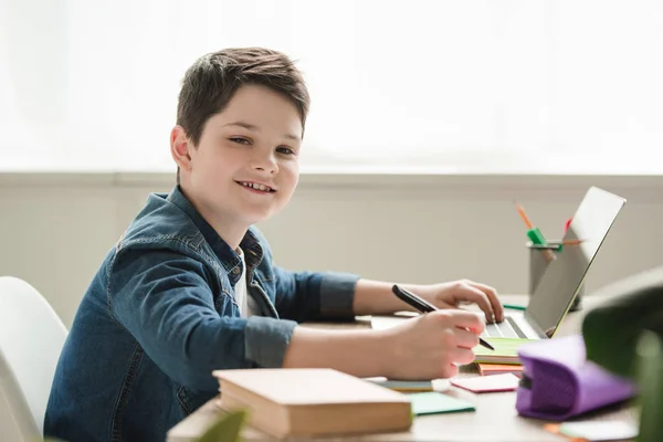 Fröhlicher Junge blickt in die Kamera, während er am Tisch sitzt und Hausaufgaben macht — Stockfoto