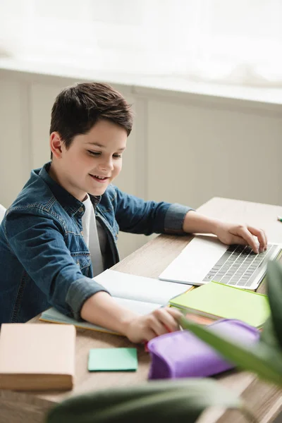 Garçon gai en utilisant un ordinateur portable tout en faisant des travaux scolaires à la maison — Photo de stock