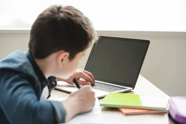 Colegial escribiendo en el cuaderno y usando el ordenador portátil mientras que hace el trabajo escolar en el país - foto de stock