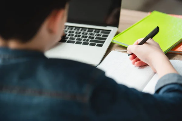 Back view of boy writing in notebook and using laptop while doing homework — Stock Photo