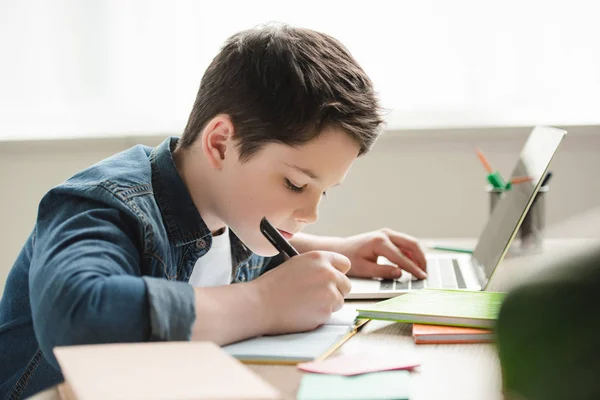 Adorable garçon attentif écrit dans un cahier et en utilisant un ordinateur portable tout en faisant des devoirs — Photo de stock