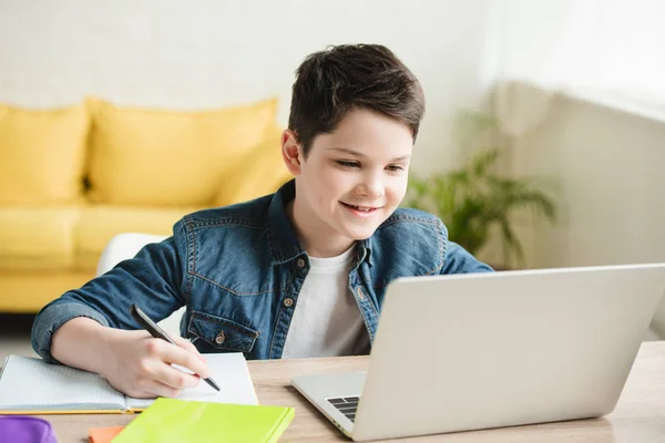 Niño alegre escribiendo en el cuaderno y usando el ordenador portátil mientras que hace el trabajo escolar en el país - foto de stock