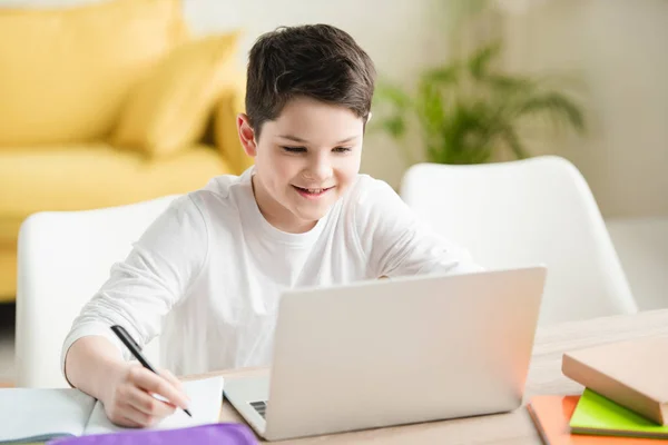 Niño alegre usando el ordenador portátil y la escritura en el libro de copia mientras que hace el trabajo escolar en casa - foto de stock