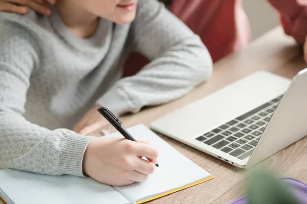 Vista parcial de niño escribiendo en el cuaderno y usando el ordenador portátil cerca de la madre - foto de stock