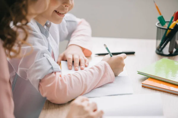 Vista recortada de la madre y la hija sentadas en la mesa y haciendo el trabajo escolar juntos en casa - foto de stock