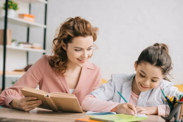 Bella donna che tiene libro e adorabile figlia che scrive nel taccuino mentre fa i compiti insieme — Foto stock