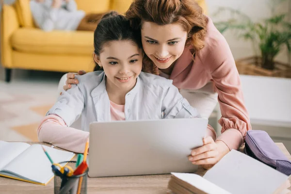 Happy mother embracing smiling daughter doing schoolwork at home — Stock Photo