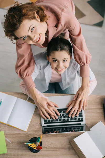 Overhead-Ansicht glücklicher Mutter und Tochter, die gemeinsam Laptop benutzen und in die Kamera schauen — Stockfoto